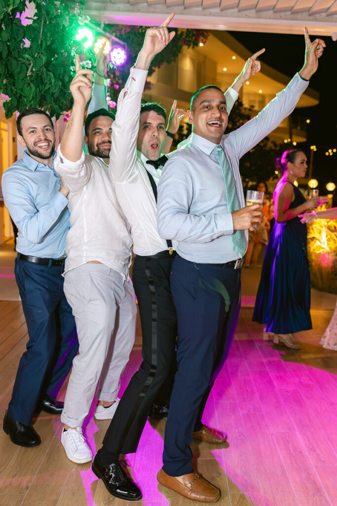 Five men in formal wear posing with raised hands at an indoor party with colorful lights.