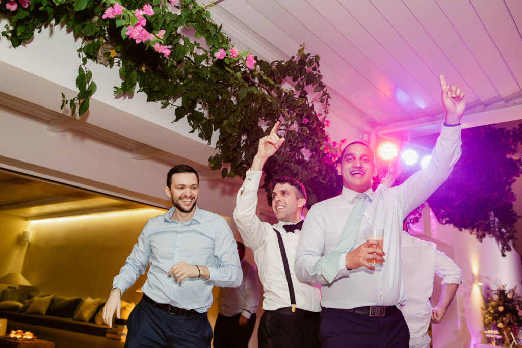 Men dancing indoors under ceiling decorated with green foliage and pink flowers with colorful lighting.