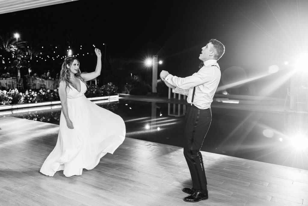 Bride in flowing gown and groom in formalwear dancing outdoors at night near a pool with lights.
