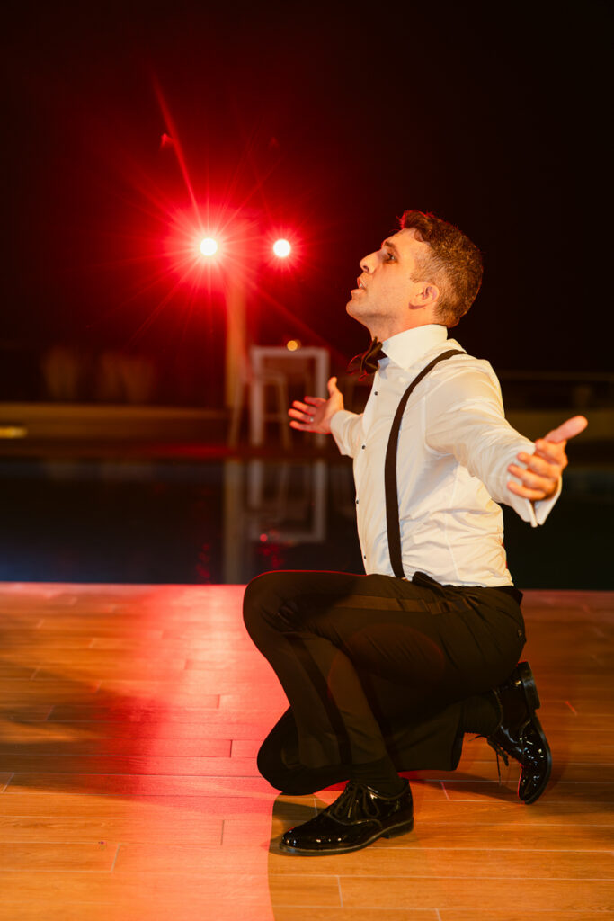 Man in white shirt with suspenders kneeling on wooden floor with arms outstretched under red lights.