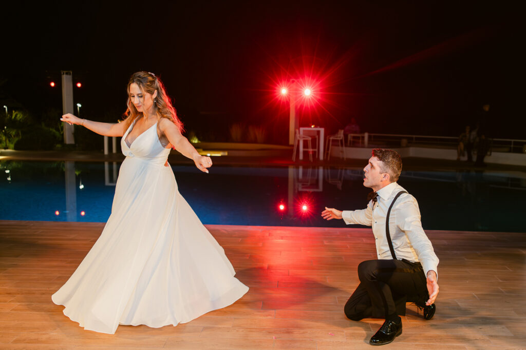 Woman in white dress dancing on wooden floor at poolside at night with man kneeling nearby.
