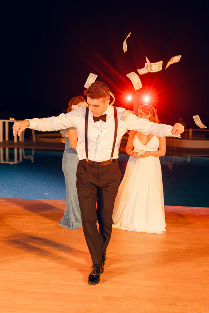 Man in formal wear dancing with flying money near two women by a pool at night.