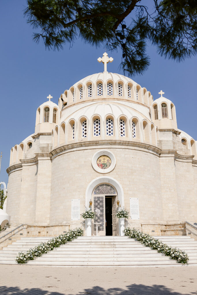 White stone church with three domes topped by crosses, ornate entrance, and floral stair decorations.