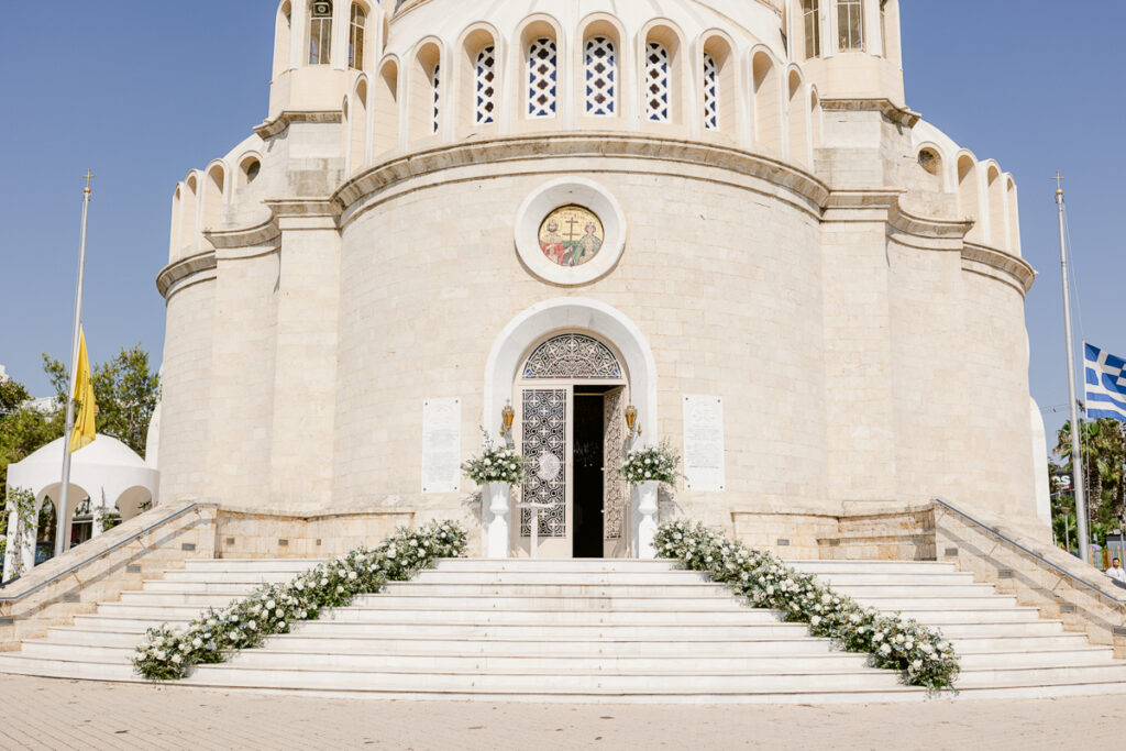 Entrance of a light stone church with floral-decorated steps, metal door, mosaic, and two flagpoles.