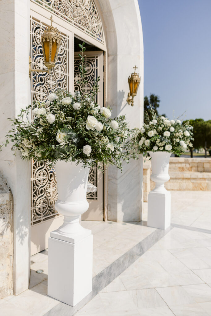 Two white urns with white flowers and greenery on white pedestals outside marble building.