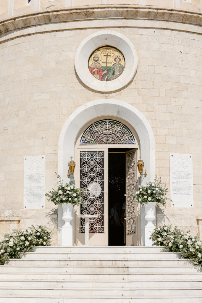 Stone church entrance with ornate wrought iron doors, mosaic above, and white floral decorations.