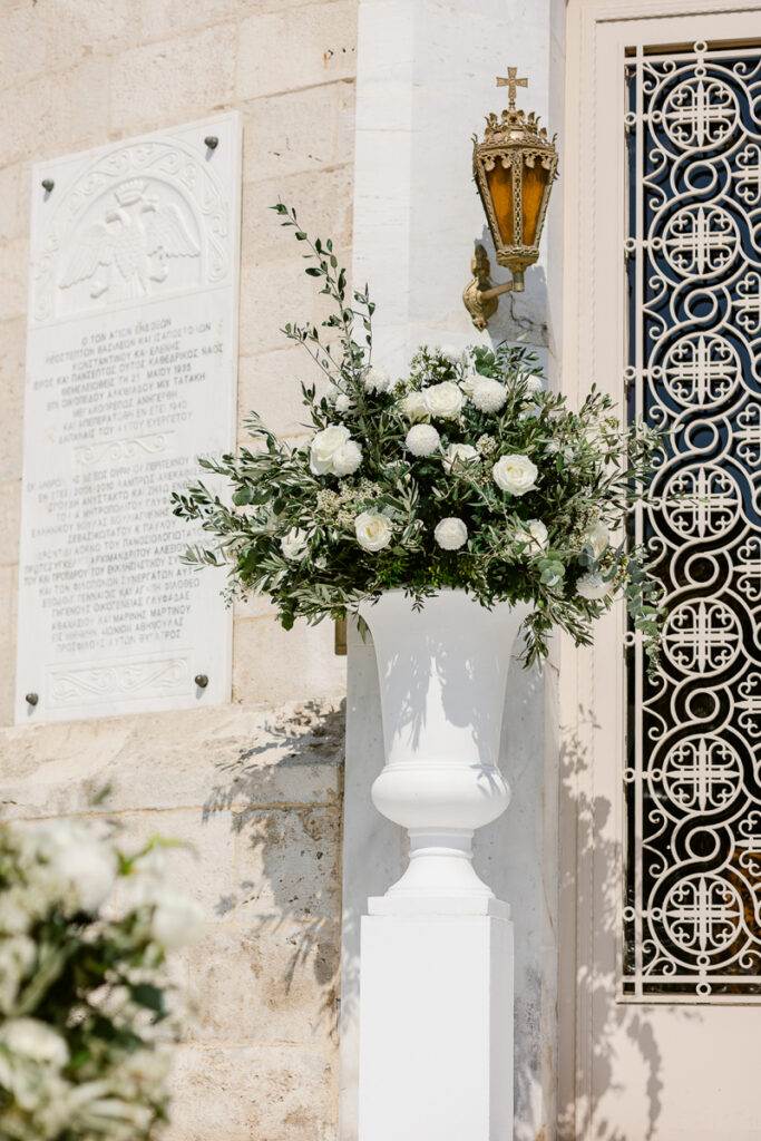 White floral arrangement in a white urn beside a stone wall plaque and ornate door with lantern above.