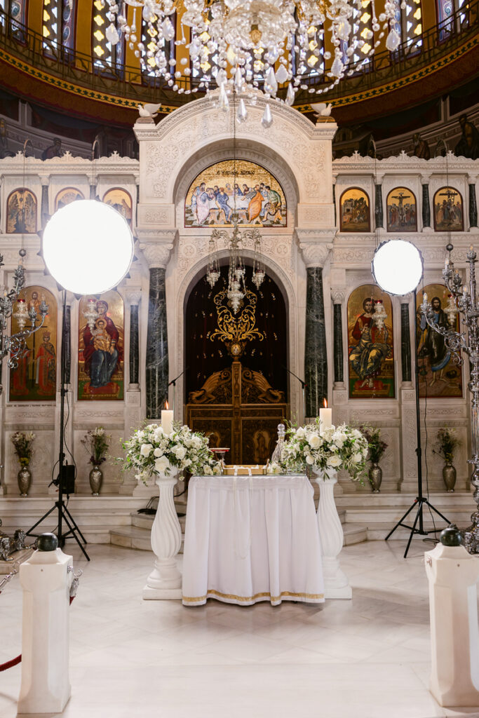 Interior of ornate church altar decorated with white flowers, candles, religious icons, and studio lights.