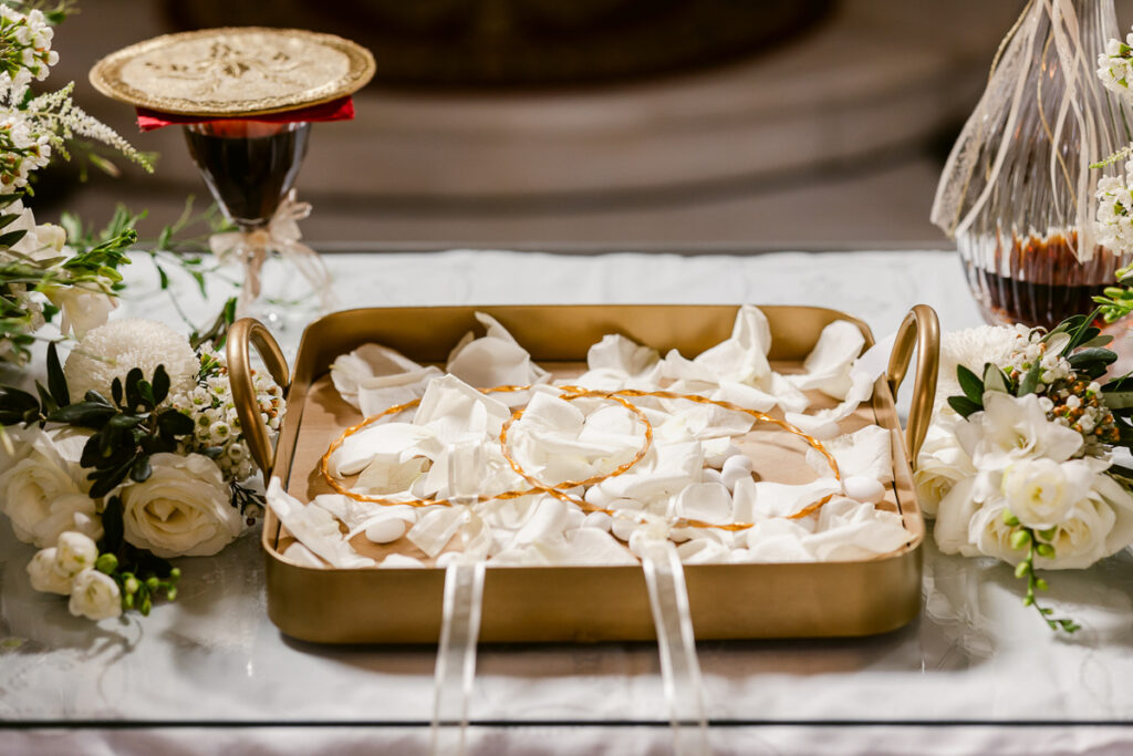 Gold tray with two intertwined gold wedding crowns and white petals on a decorated table.