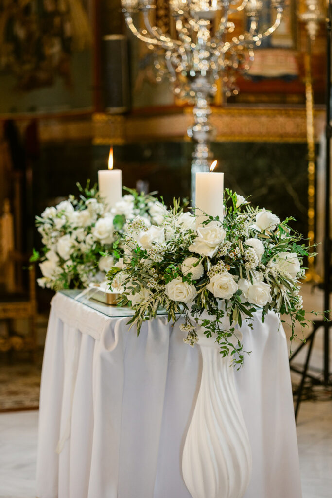 White table with floral arrangements of white roses and greenery, each holding a lit candle.