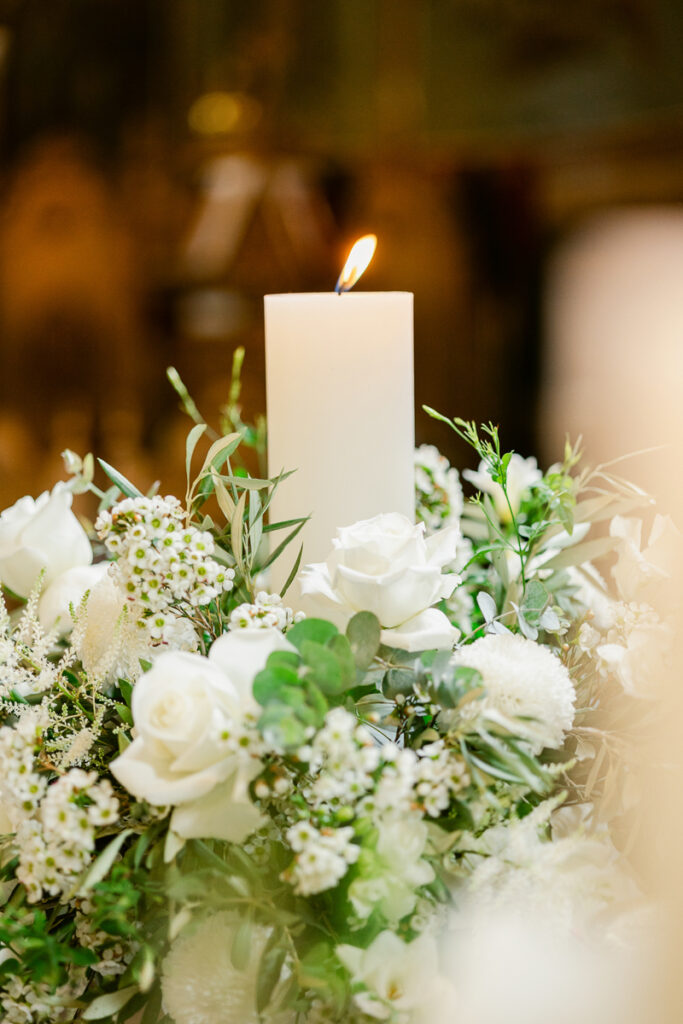 White lit candle surrounded by white roses and green foliage floral arrangement.