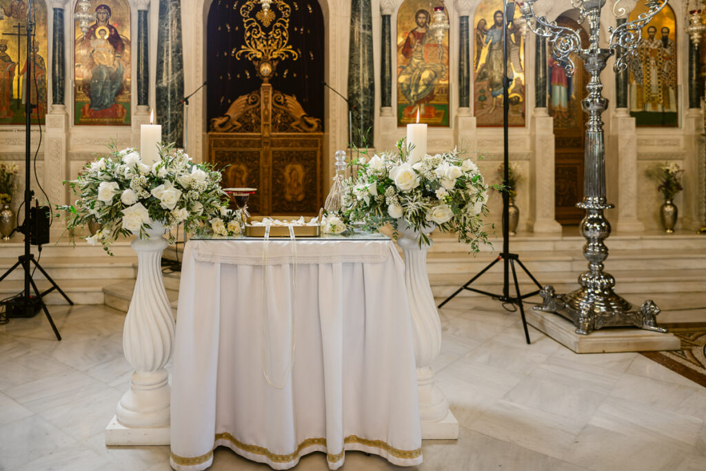 Ceremonial church table with white cloth, candles, floral arrangements, chalice, and religious icons backdrop.