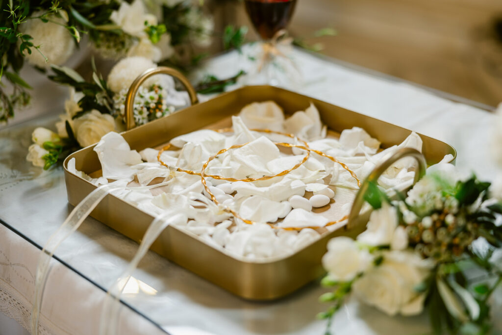 Gold tray with white rose petals, two gold rings, and ribbon on lace-covered table with flowers.