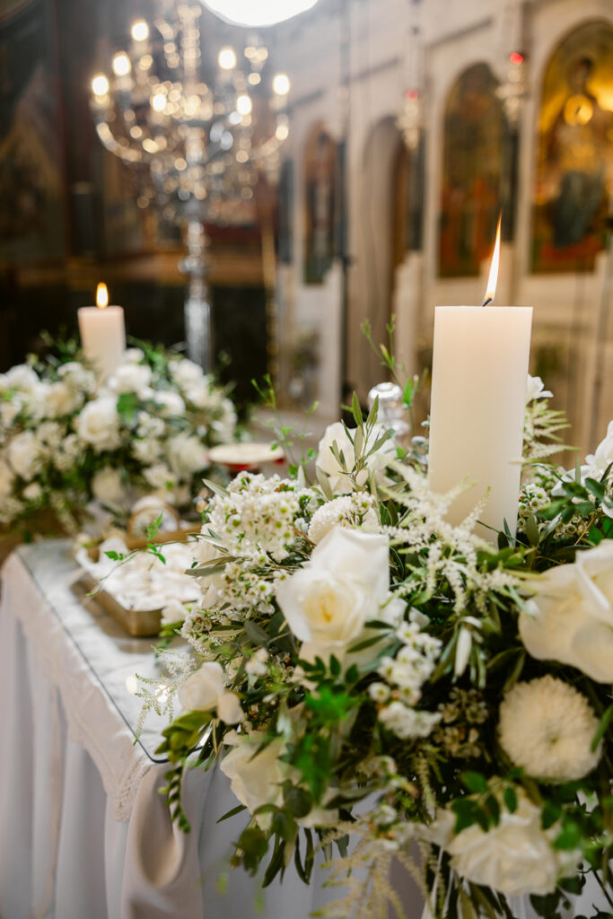 Lit white candles surrounded by white floral arrangements on a table in a decorated interior.