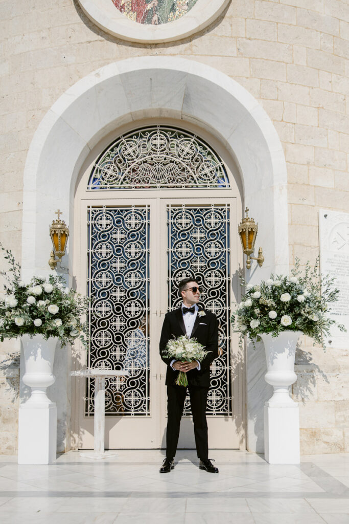 Man in black tuxedo holding white flower bouquet standing between two large white floral arrangements by ornate arched gate.