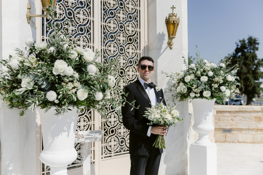 Man in black tuxedo with sunglasses holding white flower bouquet by ornate gate outdoors.