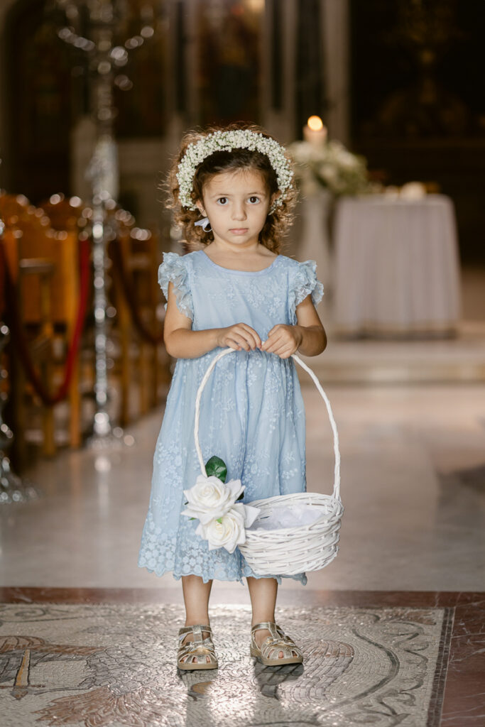 Young girl in blue lace dress and floral headband holding white wicker basket indoors.