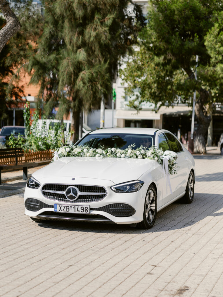 White Mercedes-Benz car decorated with white flowers and greenery on city street with trees.