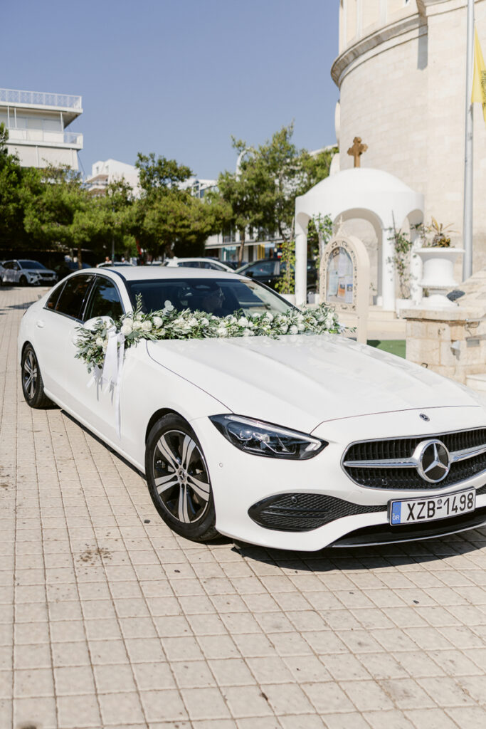 White Mercedes-Benz sedan with floral decorations parked near a small chapel and cross.