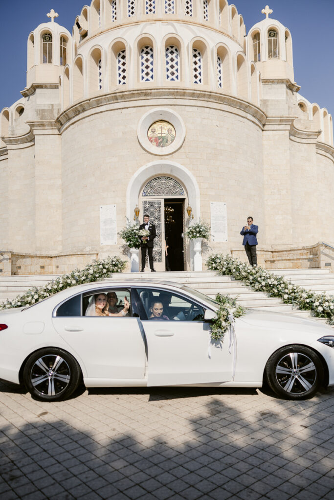 White wedding car with floral decorations parked outside a decorated stone church with people visible.