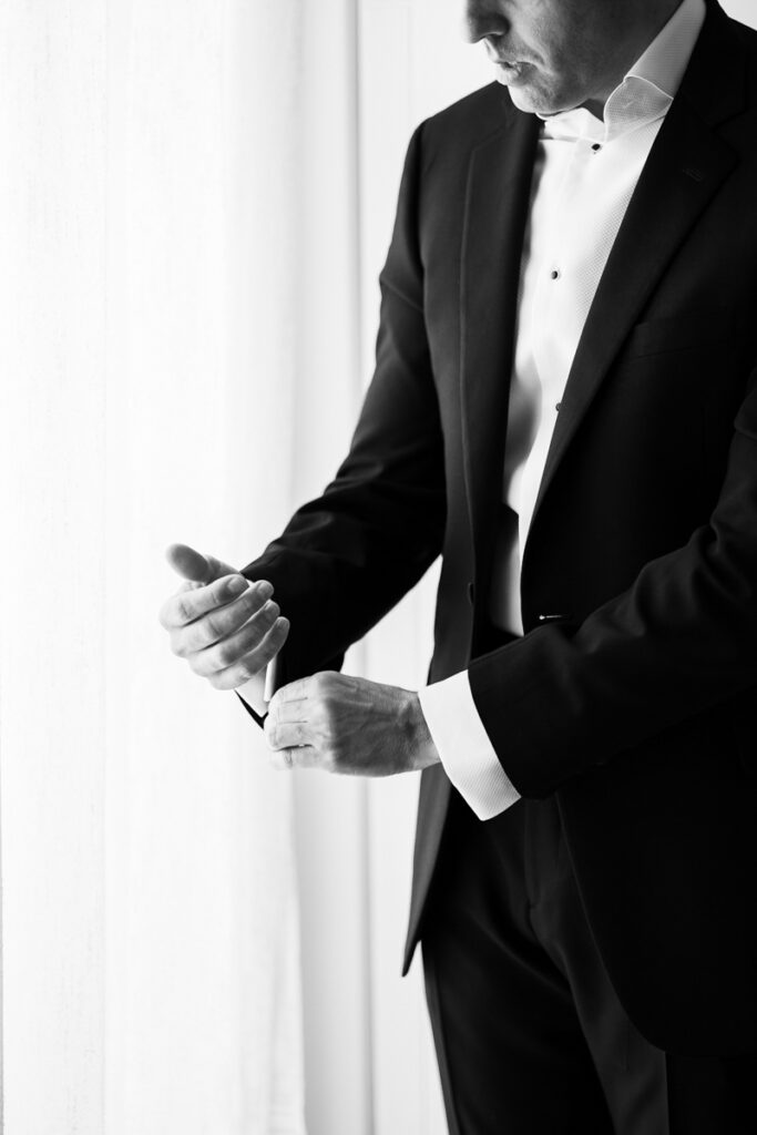 Man in tuxedo adjusting cufflinks near a window with sheer curtains in black and white.