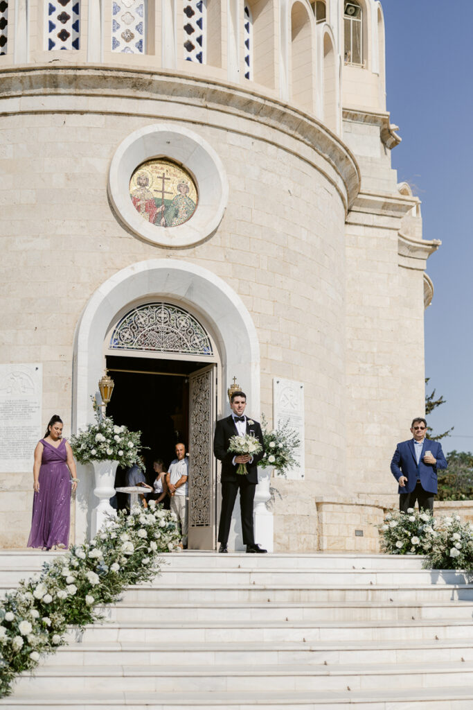 Man in tuxedo holding bouquet stands with two others on steps outside stone church entrance decorated with flowers.