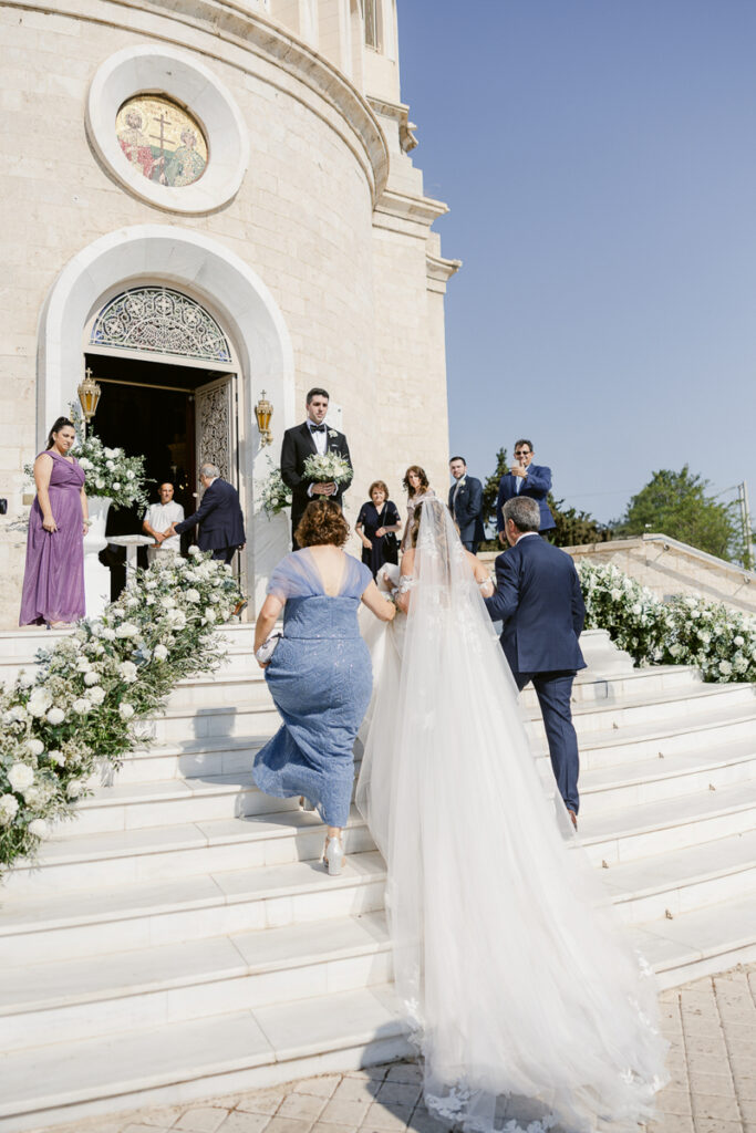 Bride in white gown ascending church steps with two attendants and guests nearby.