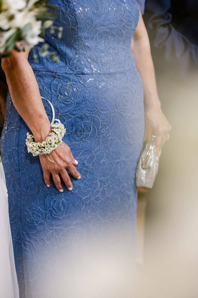 Person in blue floral lace dress with white flower corsage and holding a silver clutch.
