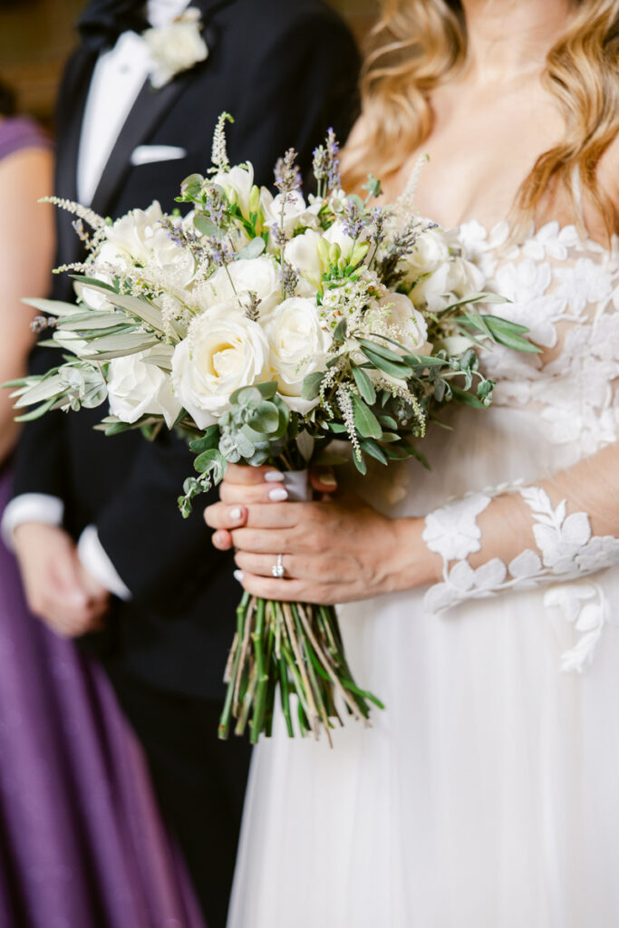 Bride in floral lace dress holding white flower bouquet with people in formal attire nearby