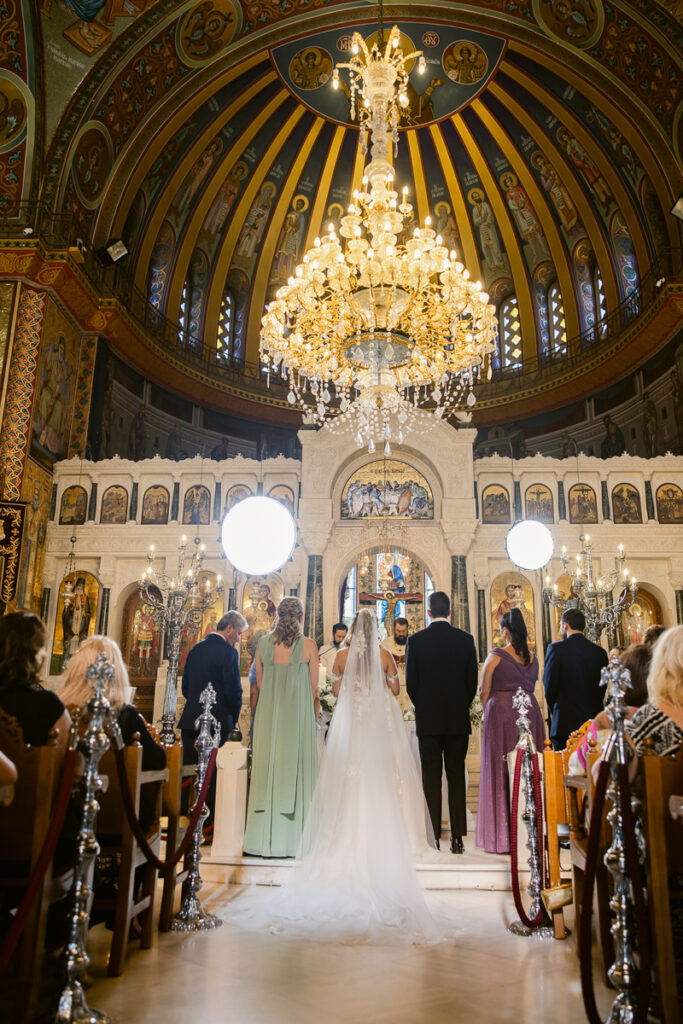 Wedding ceremony inside a decorated church with bride, groom, attendants, and guests under a chandelier.