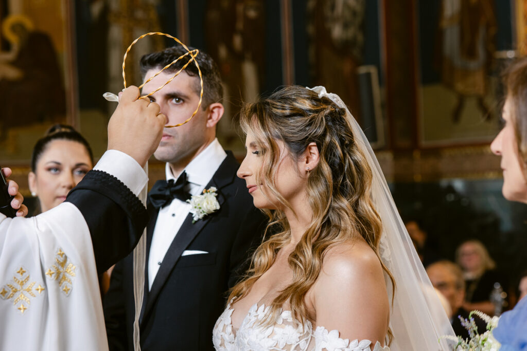 Bride in lace dress and groom in tuxedo at church wedding with priest holding braided crowns.
