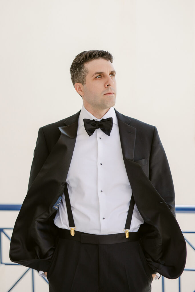 Man wearing black tuxedo jacket over shoulders, white dress shirt, black bow tie, and suspenders.