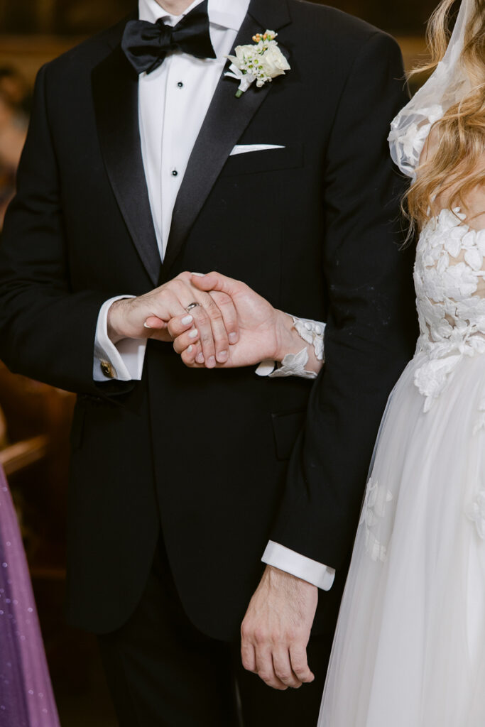 Close-up of a couple holding hands with wedding rings, dressed in wedding attire.