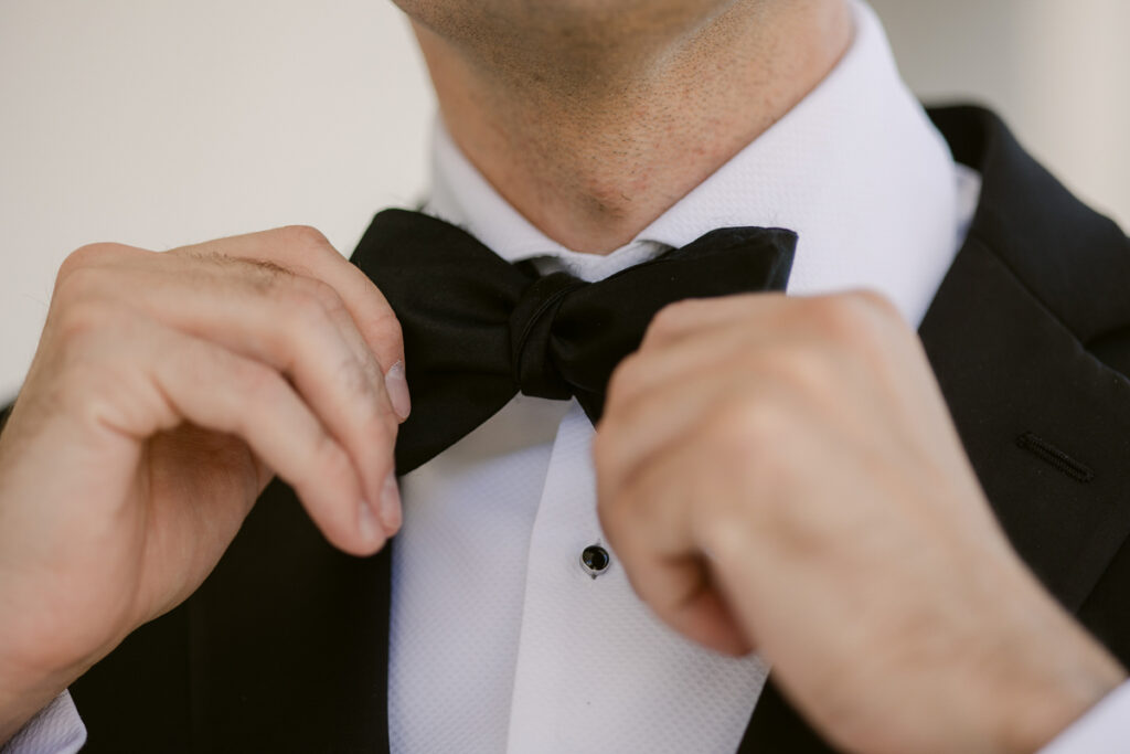 Person adjusting black bow tie on white dress shirt and black suit jacket close-up.