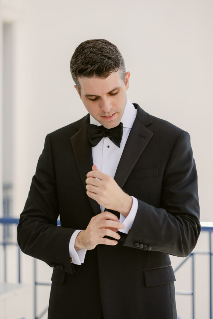 Man in black tuxedo adjusting his cufflink indoors near blue railing with light walls.