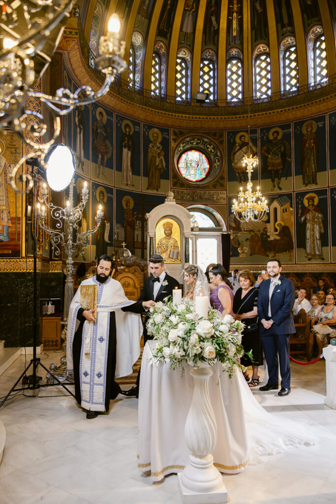 Wedding ceremony in a decorated church with bride, groom, priest, attendants, and guests.