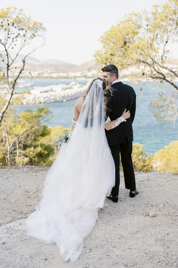 Bride in white gown and groom in suit embrace overlooking water and rocky shoreline.