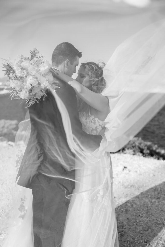 Black and white photo of bride and groom embracing near water with veil flowing in wind.