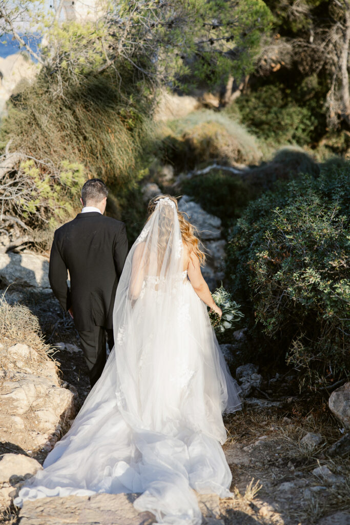Bride in white gown and groom in black suit walking on rocky outdoor path surrounded by bushes.