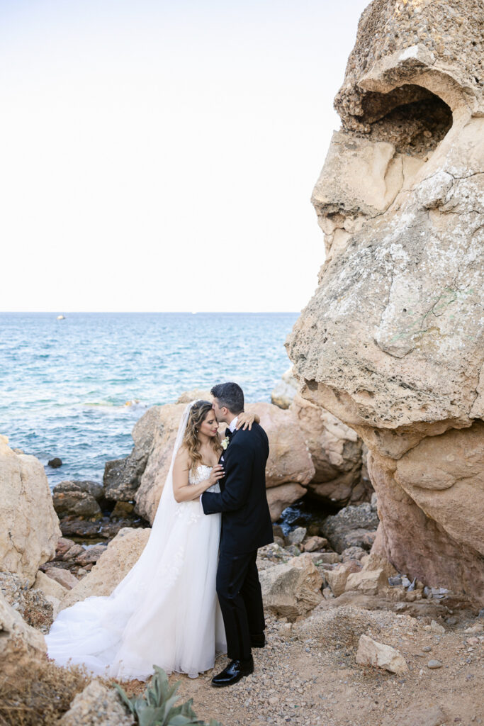 Bride in white dress and groom in black suit embrace on rocky shoreline by ocean.