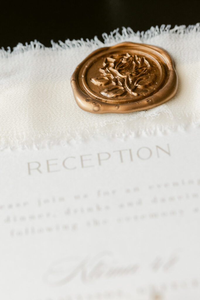 Close-up of a textured paper reception invitation with a gold floral wax seal on fabric.