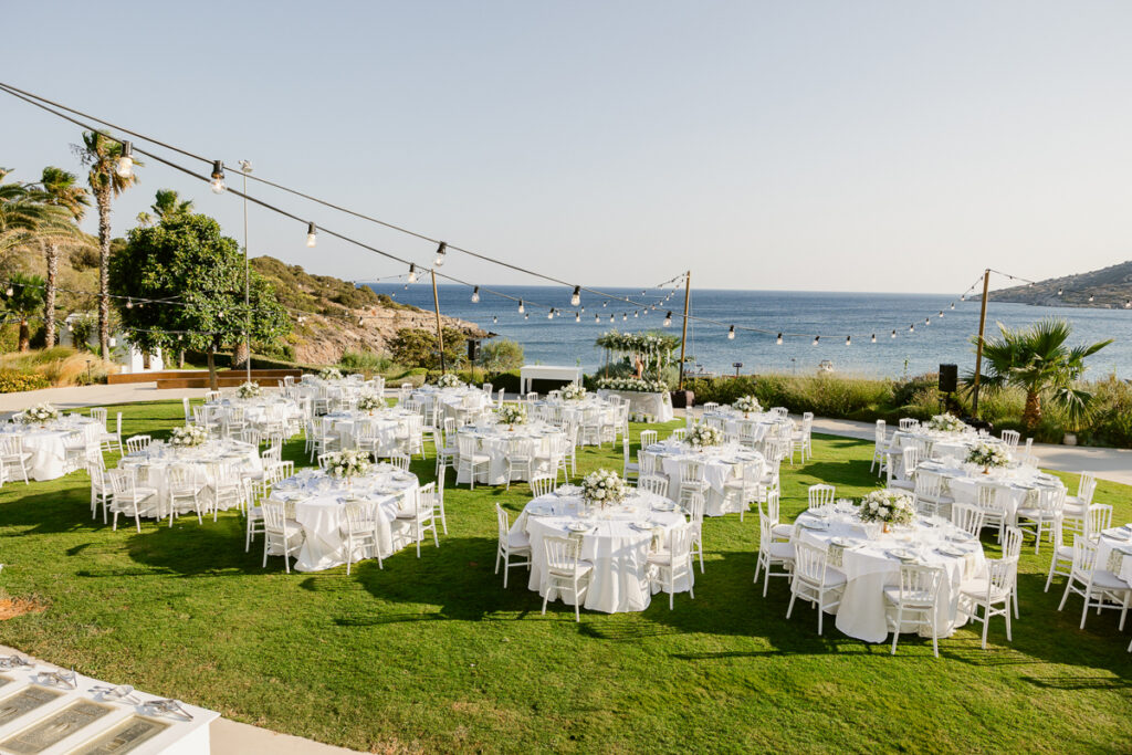 Outdoor coastal event setup with round tables, white chairs, floral centerpieces, and string lights.