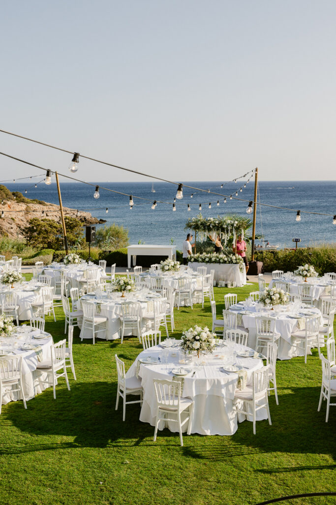 Outdoor seaside event setup with round tables, white chairs, floral centerpieces, and string lights.