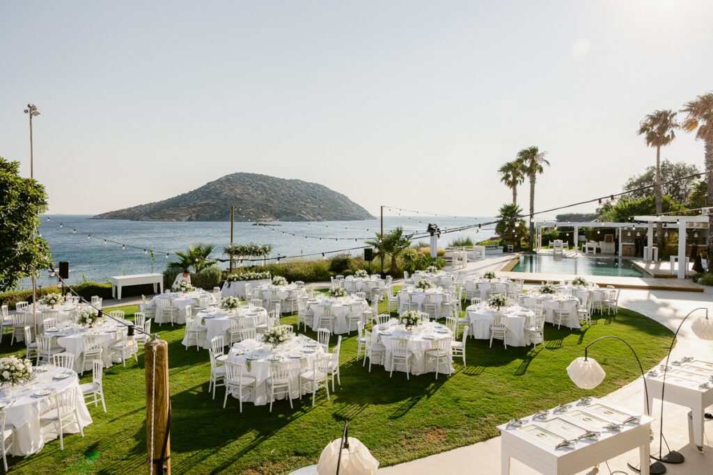 Outdoor event setup with round tables, white chairs, floral centerpieces, pool, and sea view.