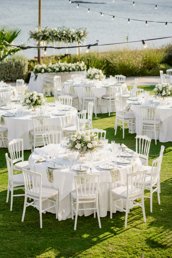 Outdoor event with round tables, white chairs, floral centerpieces, and string lights by water.