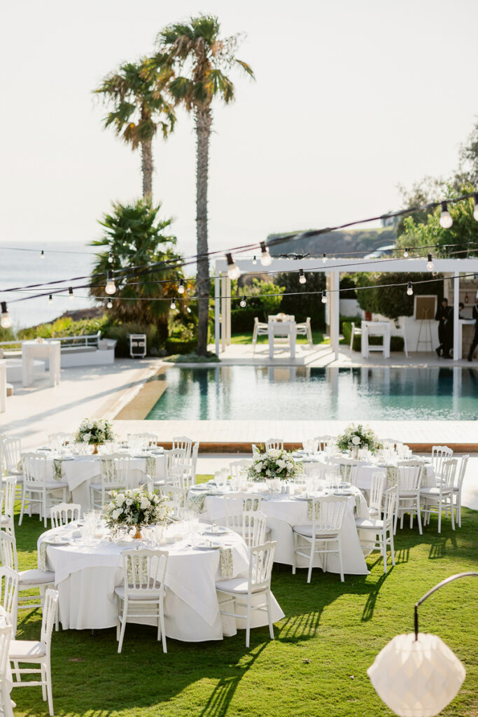 Outdoor event seating with white tables and chairs on grass near pool, palm trees, and ocean view.