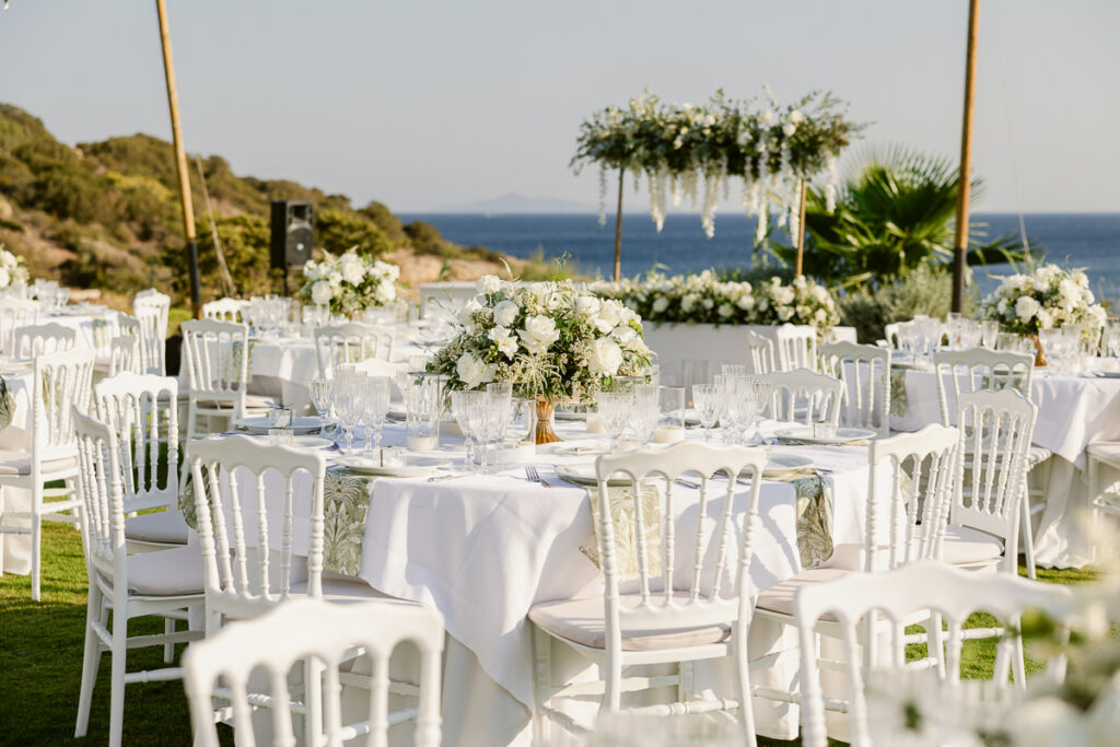 Outdoor white tablecloth dining setup with floral centerpieces near sea and hills view.