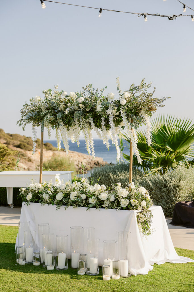 Outdoor white floral decorated table with hanging greenery and candles near coastline.