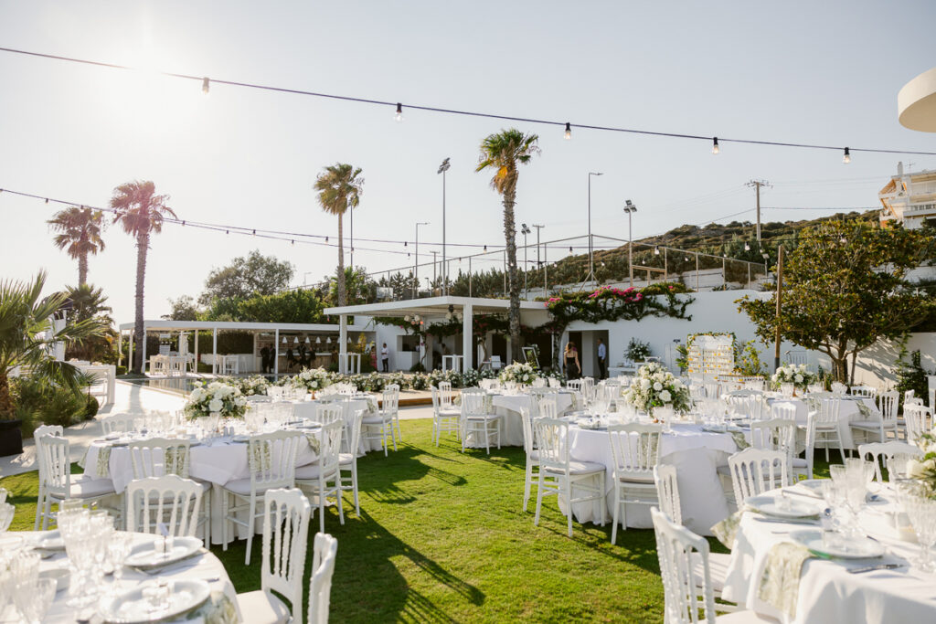 Outdoor event setup with white round tables, chairs, floral centerpieces, and string lights on green lawn.