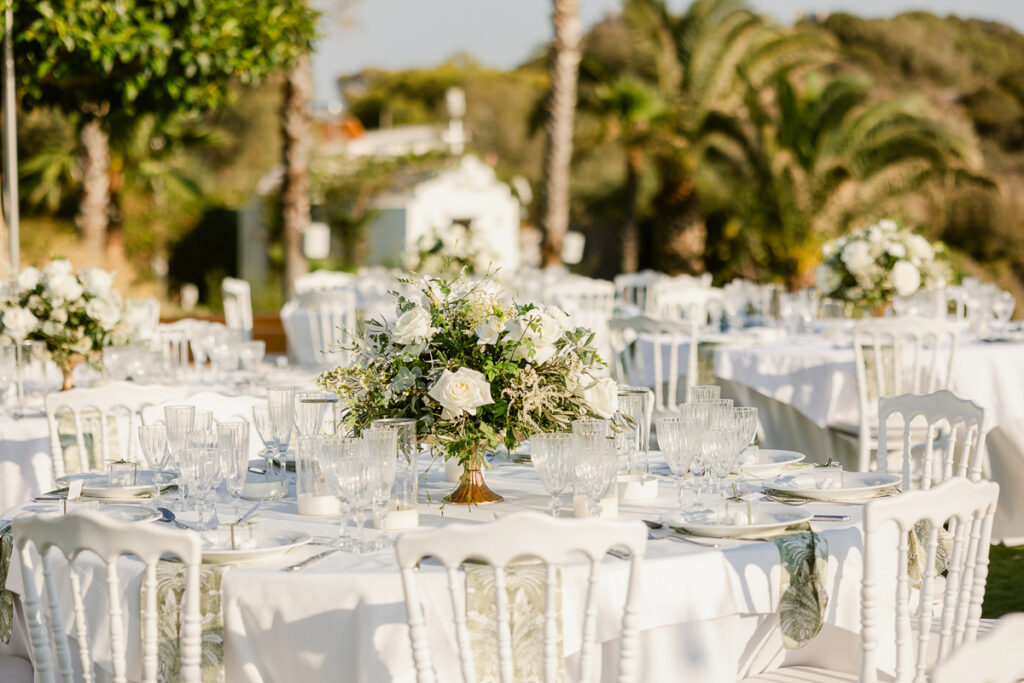 Outdoor event setup with white round tables, floral centerpieces, glassware, and white chairs.